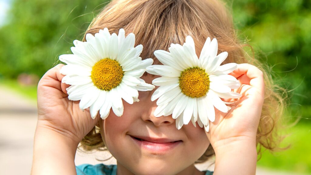 Criança sorrindo segurando duas margaridas na frente dos olhos